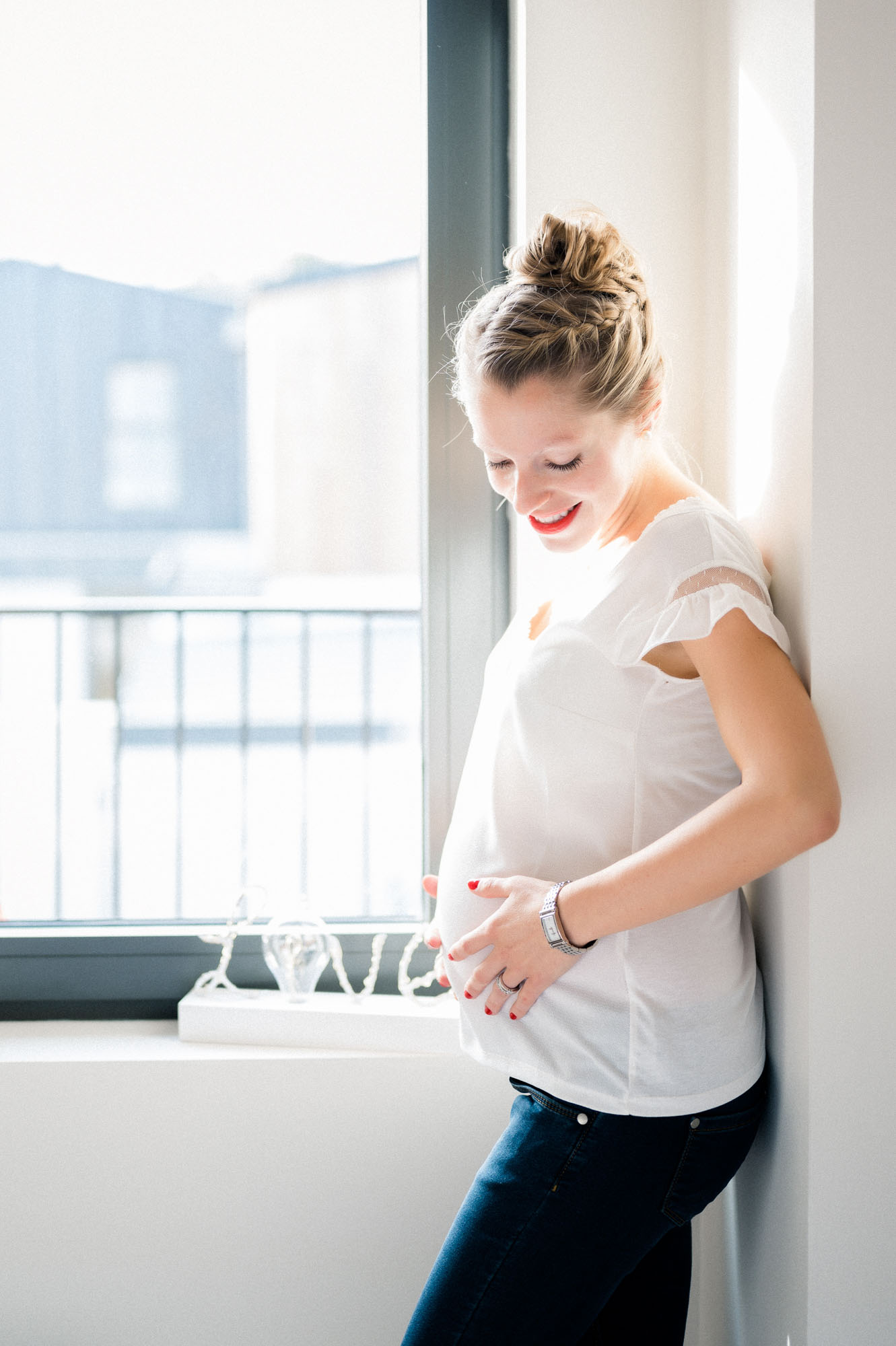 Future maman souriante tenant son ventre près d'une fenêtre, séance grossesse à Mérignac
