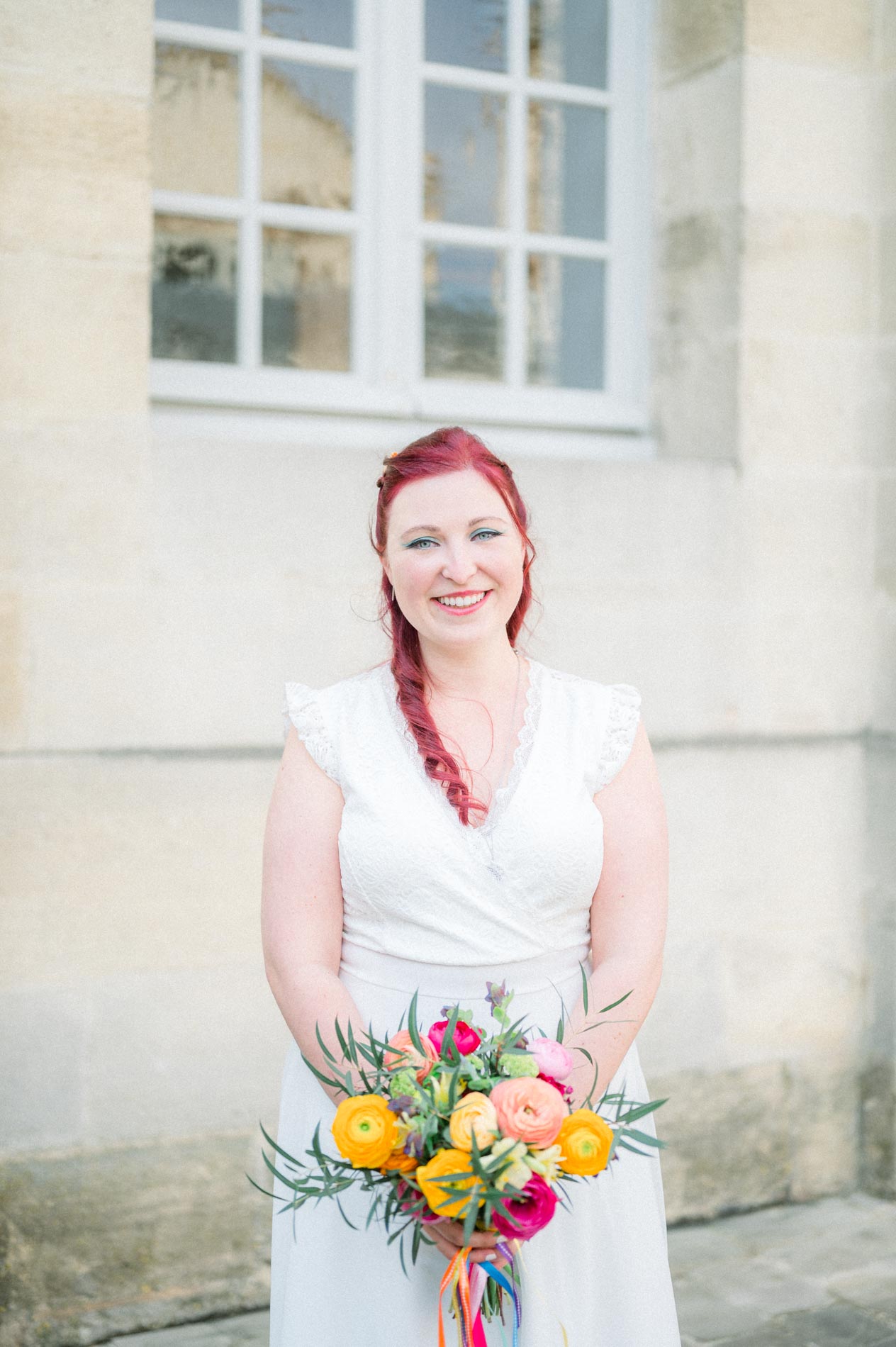 Mariée aux cheveux rouges tressés tenant un bouquet coloré devant la mairie