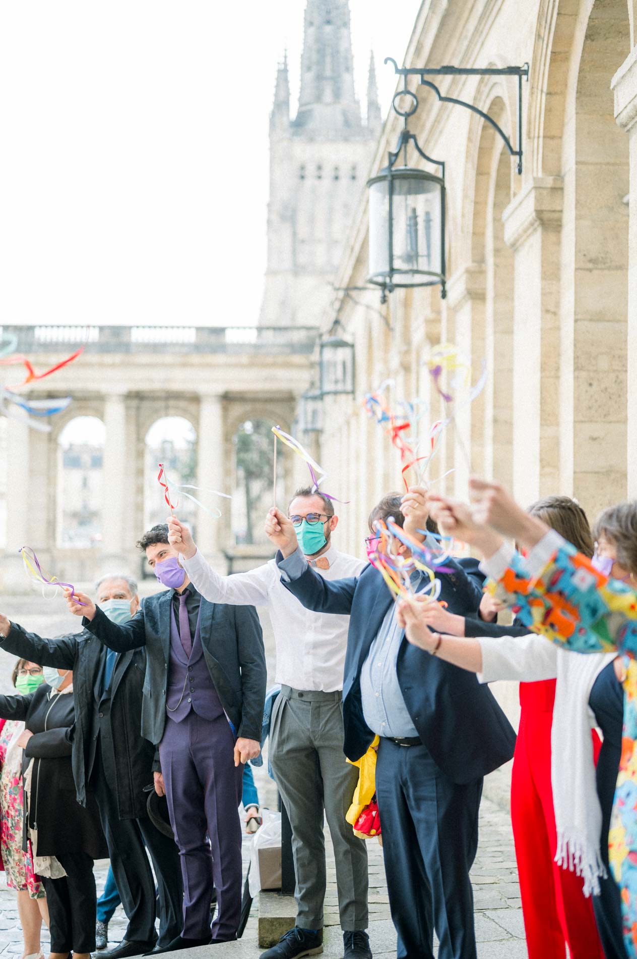 Invités agitant des rubans colorés pour la sortie des mariés sous la colonnade