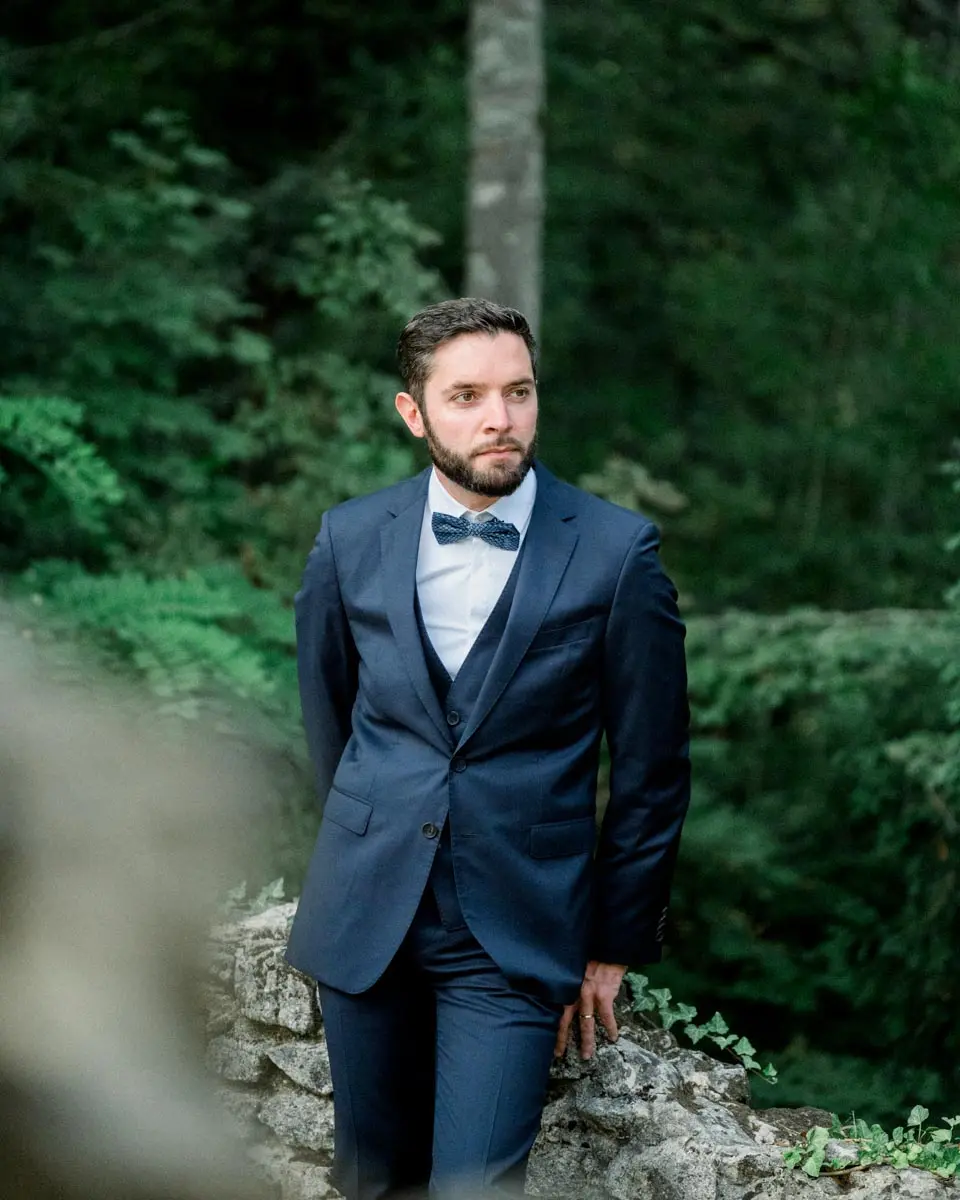Le marié en costume bleu marine adossé à un rocher dans le Parc Majolan