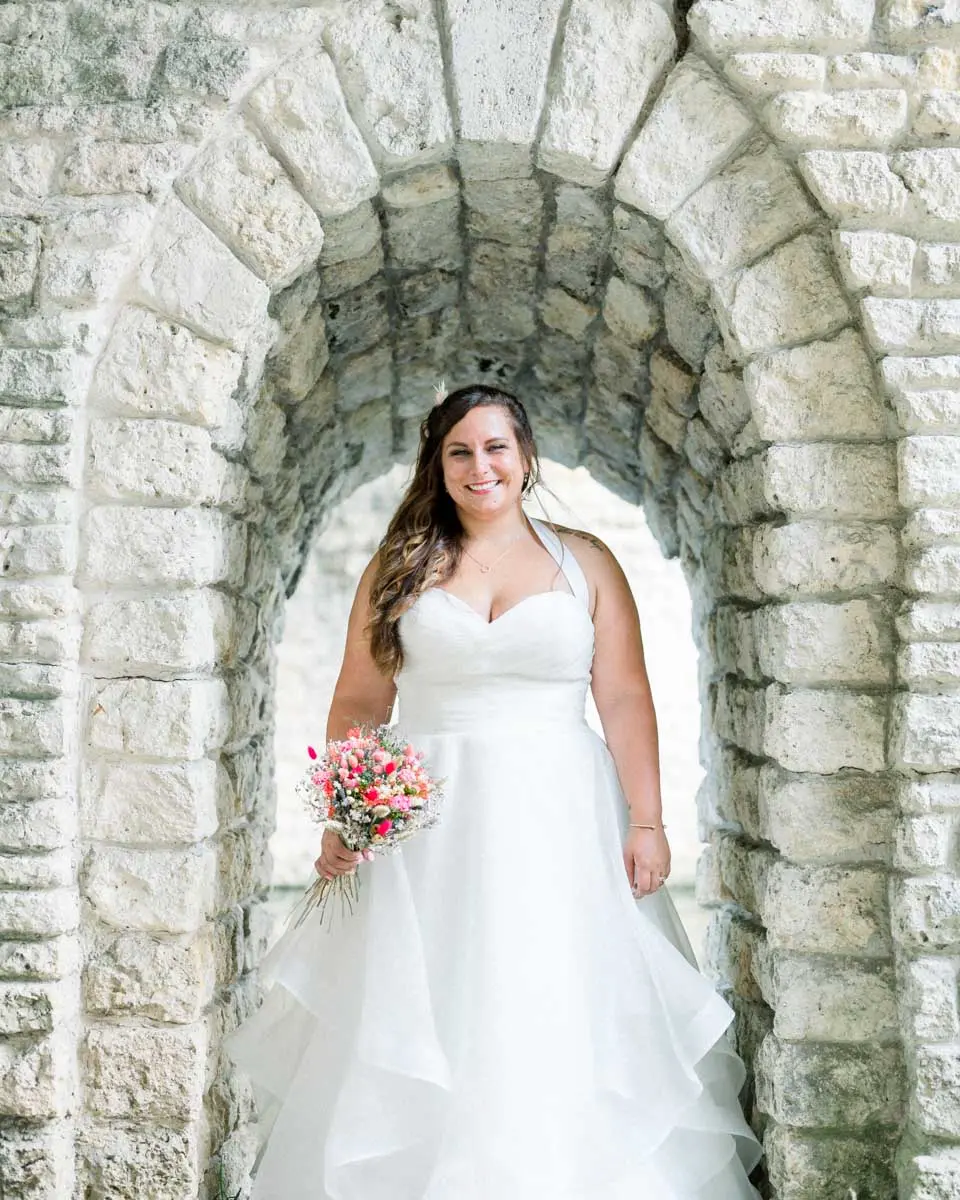La mariée sous une arche de pierre du Parc Majolan, bouquet à la main