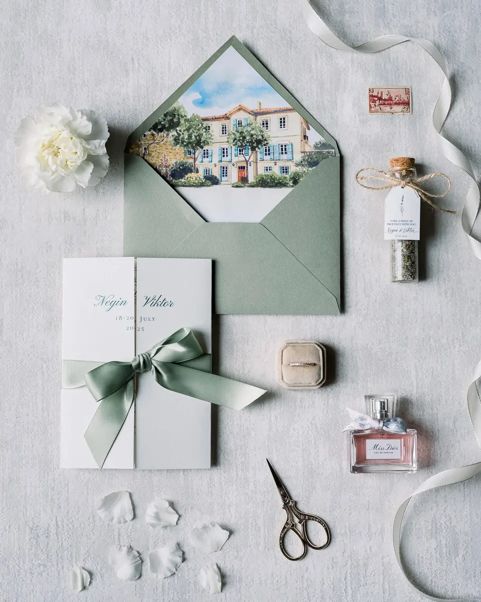 Faire-part au ruban vert sauge, écrin, parfum et ciseaux posés à plat